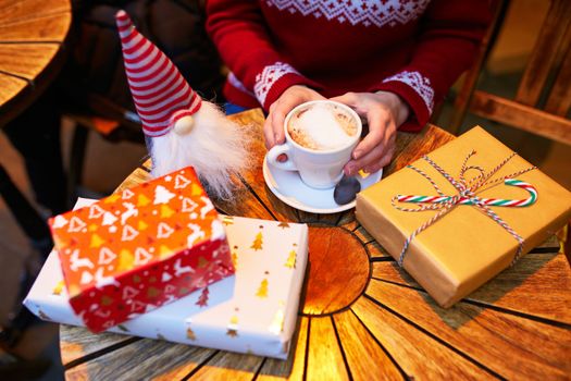 Closeup of woman's hands with cup of coffee and wrapped Christmas presents on wooden table of cafe