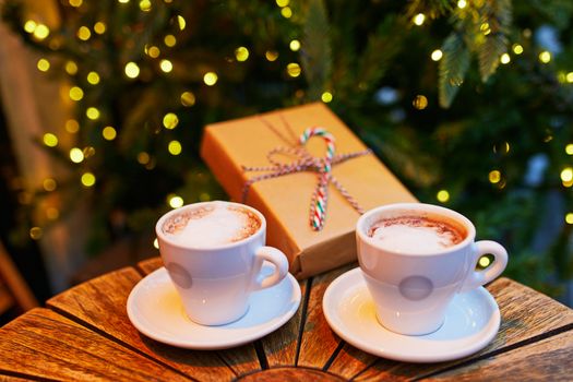 Two cups of coffee and wrapped Christmas present on a wooden table of cafe or restaurant near Christmas tree decorated with light garland