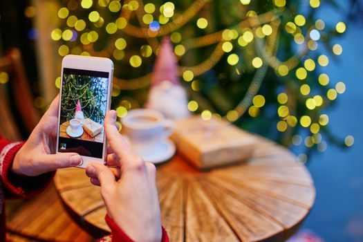 Woman taking photo of her coffee cup and Christmas present in cafe decorated for seasonal holidays