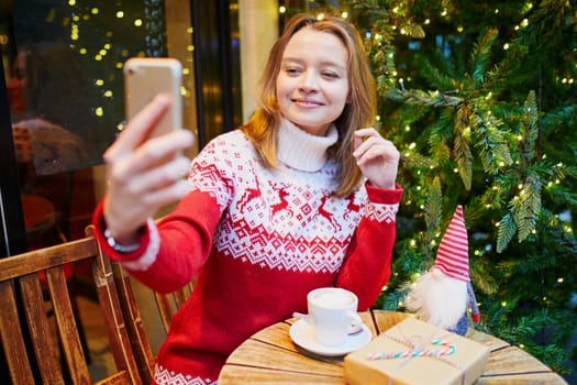 Cheerful young girl in warm red knitted holiday sweater in cafe decorated for Christmas, making selfie with her coffee cup and Christmas present