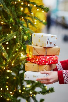 Woman hands holding pile of Christmas presents near New year tree decorated with lights and beads