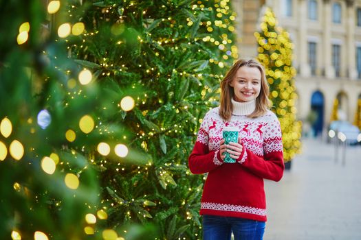 Happy young girl in warm knitted holiday sweater walking with hot drink to go on a street of Paris decorated for Christmas