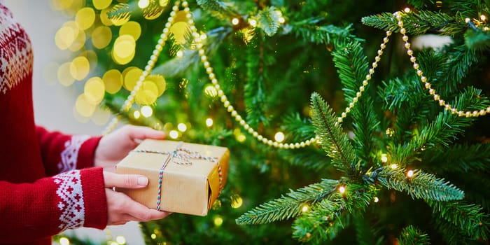 Woman hands holding Christmas present near New year tree decorated with lights and beads