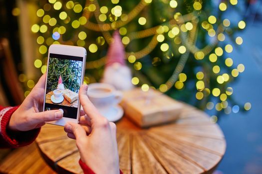 Woman taking photo pf her coffee cup and Christmas present in cafe decorated for seasonal holidays