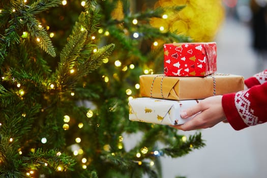 Woman hands holding pile of Christmas presents near New year tree decorated with lights and beads