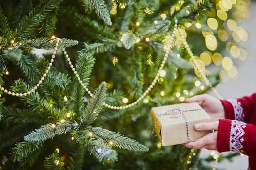 Woman hands holding Christmas present near New year tree decorated with lights and beads