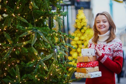 Happy young girl in warm red knitted sweater with pile of holiday gifts on a street of Paris decorated for Christmas