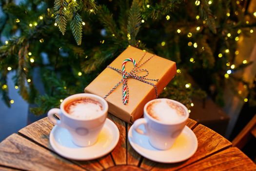 Two cups of coffee and wrapped Christmas present on a wooden table of cafe or restaurant near Christmas tree decorated with light garland