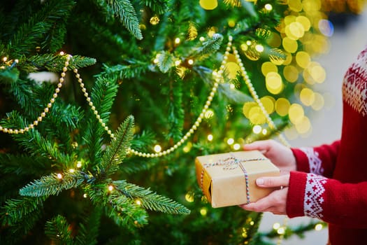 Woman hands holding Christmas present near New year tree decorated with lights and beads