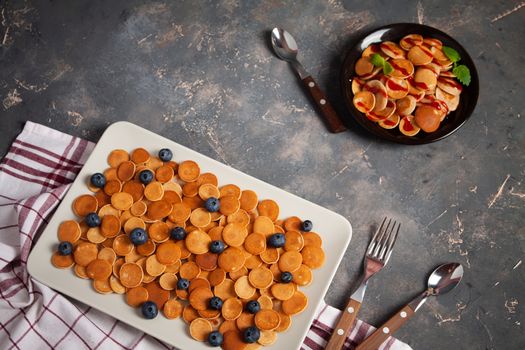 Trendy homemade breakfast, pancake cereal, mini pancakes on plates on dark concrete background with spoon and blueberry, peppermint with textile