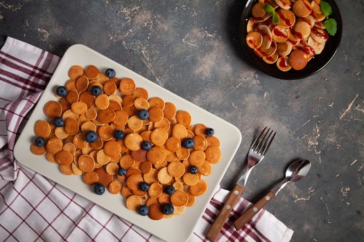 Trendy homemade breakfast, pancake cereal, mini pancakes on plates on dark concrete background with spoon and blueberry, peppermint with textile