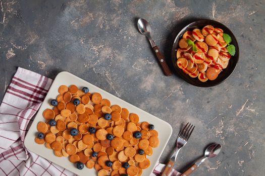 Trendy homemade breakfast, pancake cereal, mini pancakes on plates on dark concrete background with spoon and blueberry, peppermint with textile
