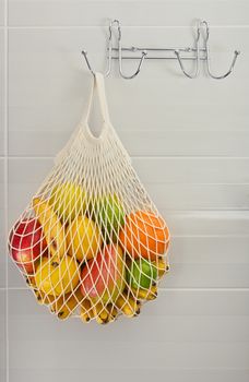 Close up assorted fresh fruits in reusable string mesh shopping bag hanging on the hook over grey wall, low angle side view