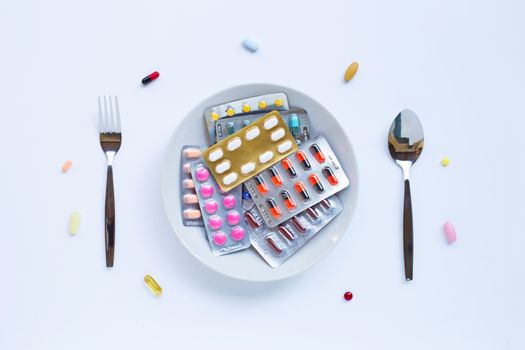Colorful tablets, capsules and pills with white dish, folk and spoon on white background.