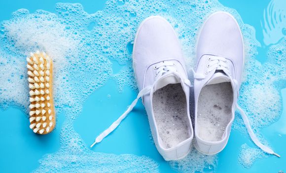 Sneakers with foam of powder detergent water dissolution and wooden brush on blue background. Washing dirty shoes. Top view