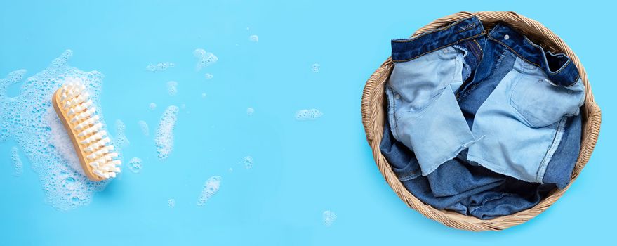 Jeans in laundry basket with wooden brush for cleaning clothes on foam of powder detergent water dissolution on blue background. Top view