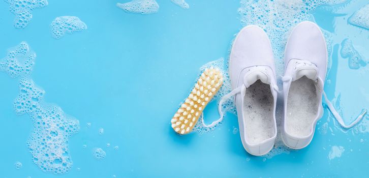 Sneakers with foam of powder detergent water dissolution and wooden brush on blue background. Washing dirty shoes. Top view