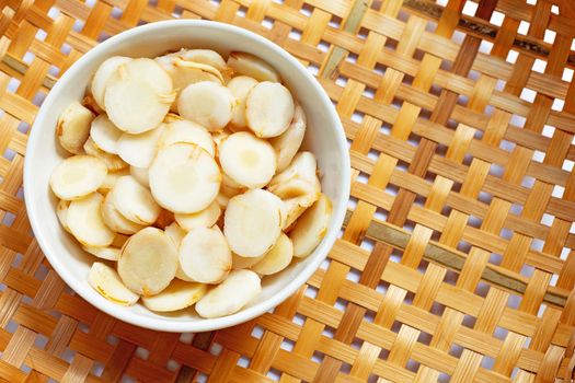Fresh galangal slices in white bowl on wooden bamboo threshing basket background. Top view