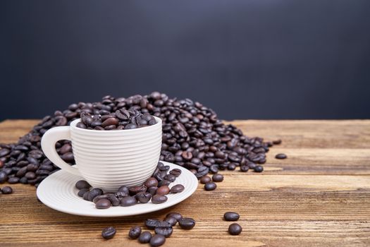 Close up coffee bean in white cup with saucer have blur pile of coffee bean overflow on old wooden table with black background and copy space.