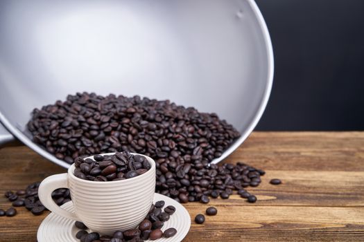 Close up coffee bean in white cup with saucer have blur pile of coffee bean in pan overflow on old wooden table with black background and copy space.