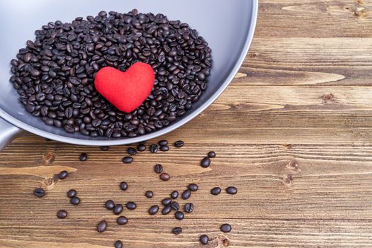 Close up red heart on pile of coffee bean in iron pan with spread coffee bean on old wooden table and copy space.