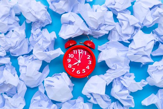 Red alarm clock with white crumpled paper balls on a blue background. Top view