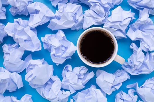 Coffee cup with white crumpled paper balls on a blue background. Top view