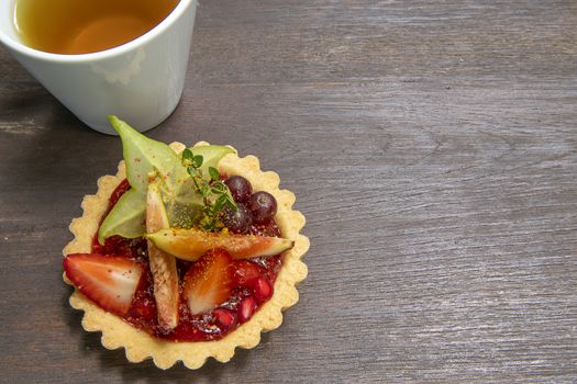 Mixed fruits tart with fig, pomegranate, starfruit, cherry, blueberry, grape, and raspberry jam. A cup of tea, brown wooden background, top view.