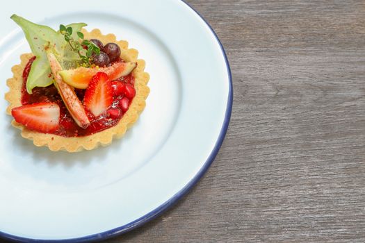 Mixed fruits tart with fig, pomegranate, starfruit, cherry, blueberry, grape, and raspberry jam on a white tin plate, brown wooden background, top view.