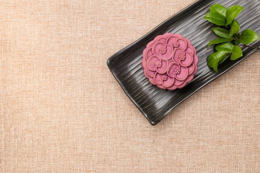 Purple japanese yam moon cake on a black ceramic plate and a twig of tea leaves. Top view, light brown cloth background