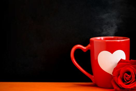 A red ceramic coffee mug with a white heart piant and a blooming red rose on an orange office desk, dark background.