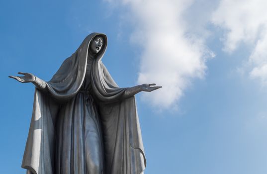 Statue of Virgin Mary in bronze, in a blue sky background with white clouds, with outstretched arms.