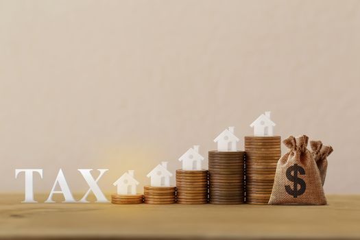 Mini house on rows of rising coins, US dollar bags, word tax on wood table. House or housing, land value, finance tax concept: depicts ad valorem tax on the value of the property all.