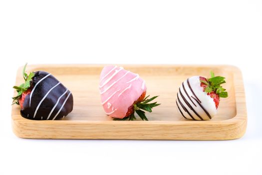 three chocolate coated strawberries (dark, pink and white) on a wooden tray, isolated on white background