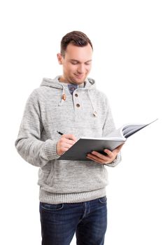 Smiling handsome male student in casual clothes, standing and writing in a notebook, isolated on white background. Young man writing in a notebook. Education concept.