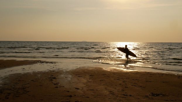Silhouette of surf man with surfboard running on water surface. Surfing at sunset beach. Outdoor water sport adventure lifestyle.Summer activity. Handsome Asia male model in his 20s.