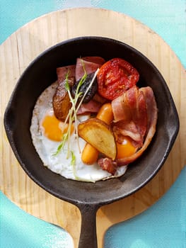 Breakfast in cooking pan with fried eggs, sausages, bacon , tomato , potato and vegetable on wood block background on the table in the kitchen. morning light effect