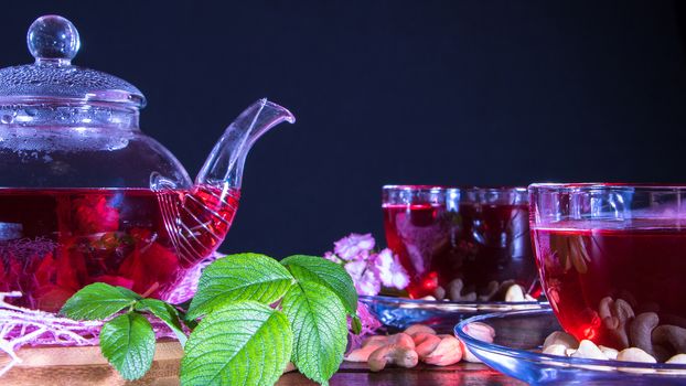 A mug of red tea and a teapot in white hibiscus flowers and green leaves of medicinal tea on a wooden stand.Zen tea ceremony. Photo of red herbal Indian healing tea.Elegant mugs with a relaxing drink