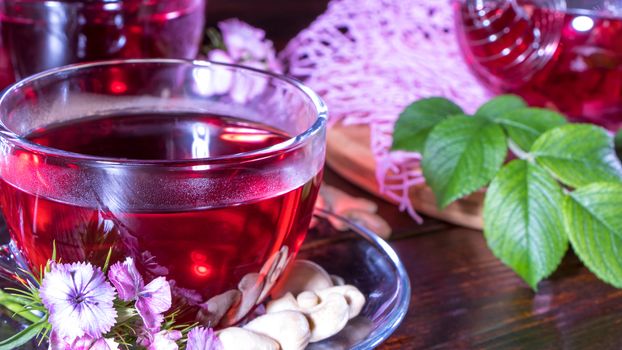 A mug of red tea in white hibiscus flowers and green leaves of medicinal tea on a wooden stand.Zen tea ceremony. Photo of red herbal Indian healing tea. Elegant mugs with a relaxing and tonic drink