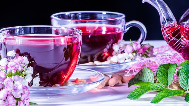 A mug of red tea in white hibiscus flowers and green leaves of medicinal tea on a wooden stand.Zen tea ceremony. Photo of red herbal Indian healing tea. Elegant mugs with a relaxing and tonic drink