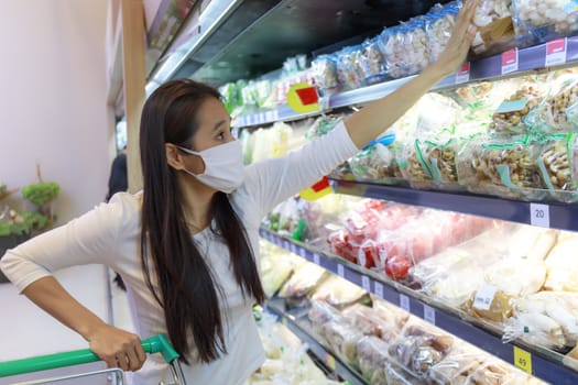 Asian woman wearing wearing protective face mask push and hold shopping cart in supermarket department store. Girl, looking grocery to buy  some food. New normal after covid-19. Family concept.