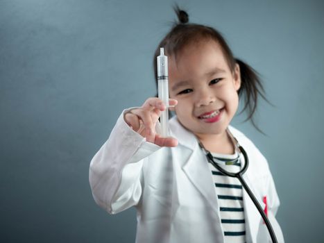 Asian little girl role playing doctor occupation wearing white gown uniform and holding a syringe. Playing is learning of children. Selective focus.