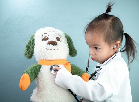 Asian little girl role playing doctor occupation wearing white gown uniform and use stethoscope to plush toy bear. Playing is learning of children.