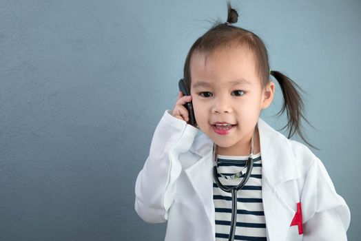 Asian little girl role playing doctor occupation wearing white gown uniform with smile and talking by phone. Playing is learning of children.