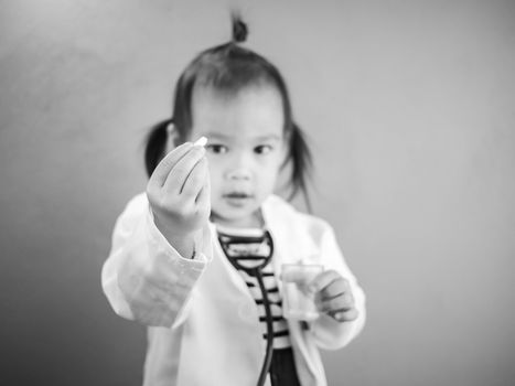 Asian little girl role playing doctor occupation wearing white gown uniform with smile and holding pill. Playing is learning of children. Selective focus.