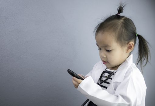 Asian little girl role playing doctor occupation wearing white gown uniform with smile and holding phone. Playing is learning of children.