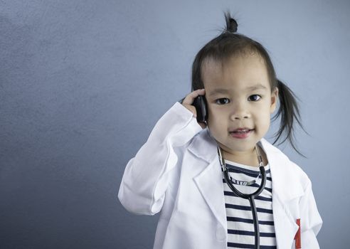 Asian little girl role playing doctor occupation wearing white gown uniform with smile and talking by phone. Playing is learning of children.