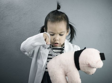 Asian little girl role playing doctor occupation wearing white gown uniform and injecting to plush toy bear. Playing is learning of children.