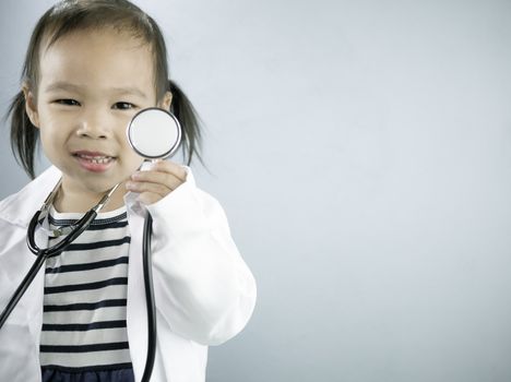 Asian little girl role playing doctor occupation wearing white gown uniform and holding a stethoscope. Playing is learning of children.