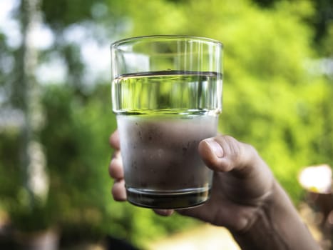 Close-up hand of woman holding cold water in clear glass over nature background.